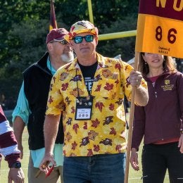 man in sunglasses, bright yellow floral shirt and boat cap, holding a guidon flag with NU-86 written on it. Several individuals follow him. Photo of guidon holder Phil Baumann '86, M'14 & P'19.