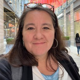 A person with dark hair stands outdoors wearing glasses on their head and hoop earrings with buildings behind them.