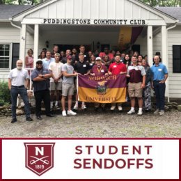 A large group of individuals stand at the entrance to a building with sign: Puddingstone Community Club.  Five individuals center front hold up a maroon and gold flag for Norwich University.