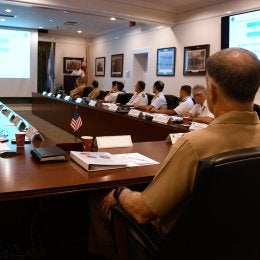 Military officers attend a conference room presentation with slides.