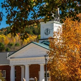 People walk past a brick building with white columns and a clock tower framed by autumn trees.