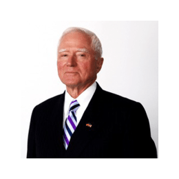 Older person with white hair wears a dark suit, striped tie, and lapel pin against a plain light background.