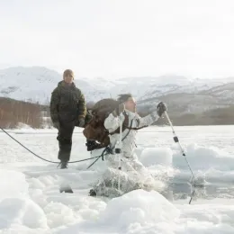 Two soldiers in black and green uniform watching another soldier in white and grey uniform jump into a hole in the ice.