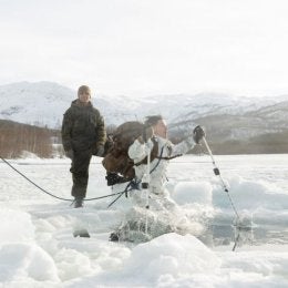 Two soldiers in black and green uniform watching another soldier in white and grey uniform jump into a hole in the ice.