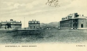 Historic black and white postcard showing three brick buildings of Norwich University campus in Northfield, Vermont, with mountains in background and open field in foreground.