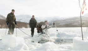 Two soldiers in black and green uniform watching another soldier in white and grey uniform jump into a hole in the ice.
