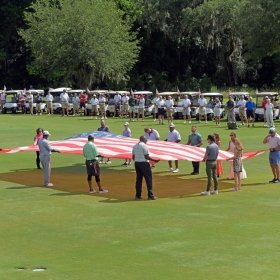 A group of people hold the American flag over a fairway green, while a line of golf carts observes from behind.