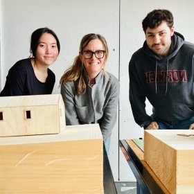 Three individuals standing behind architectural models, smiling at the camera in a workshop setting. The person on the right is wearing a Norwich University sweatshirt.