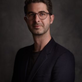 Portrait of an individual with glasses wearing a dark blazer, set against a textured grey background.