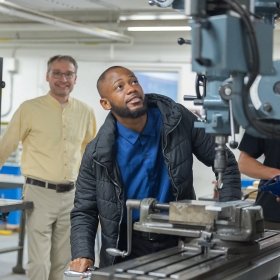 An individual operating a vertical milling machine in an industrial setting while another person observes in the background.