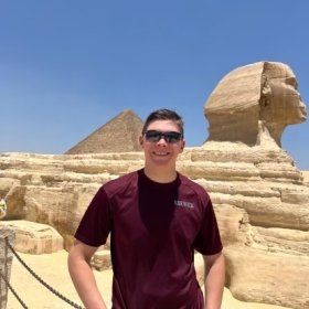Person in sunglasses and a t-shirt standing in front of the Sphinx and a pyramid in Egypt.