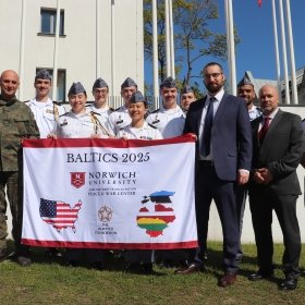A group photo of the Olmsted trip group, holding a branded Norwich/Olmsted flag in uniform in front of military academy.