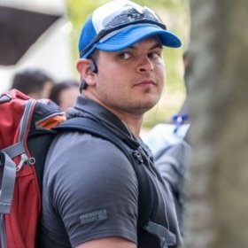 Jayden LaVecchia stands with a hat and sunglasses on top, in a polo wearing a backpack looking over his right shoulder.