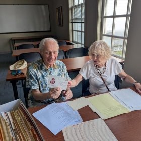 Sergey (left) in a Hawaiian styled shirt holds a letter while sitting at a table, while his wife Anna (right) points at the letter while smiling. Papers are strewn across the table.