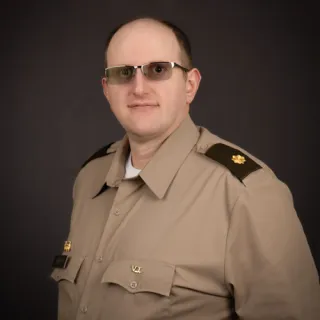 Person wearing a tan military uniform with dark shoulder epaulets and a gold oak leaf insignia, standing against a dark background.