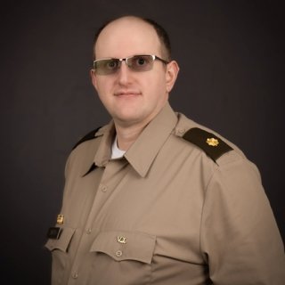 Person wearing a tan military uniform with dark shoulder epaulets and a gold oak leaf insignia, standing against a dark background.