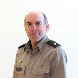 Person in a gray uniform shirt with shoulder insignia faces the camera against a plain white background.
