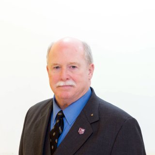Person in a dark suit, blue shirt, and patterned tie poses against a plain light background.