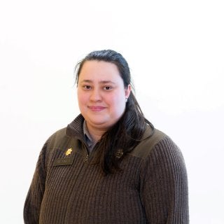 Person with long hair wears a dark sweater and faces the camera against a white background.
