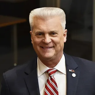 Keith Gelinas ’84: Keith Gelinas smiles for a headshot, wearing a dark suit, white shirt, and red-and-white striped tie.