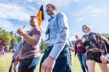 five individuals in casual clothing marching on a football field. Man leading the group holding a flag / guidon.