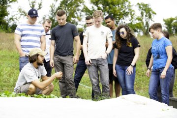 Instructor kneels in a field while a group of students stand around observing plants.