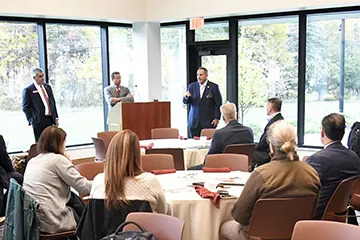 A speaker addresses attendees at a Norwich University Board of Fellows gathering while two others stand near the podium.