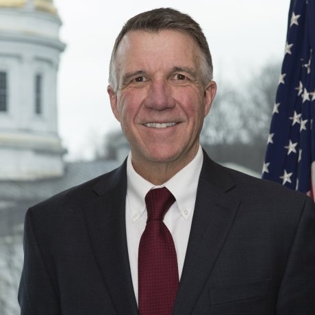 A formal headshot of Governor Phil Scott in a suit flanked by an American flag behind his left shoulder, and the capitol building in the distance behind his right.