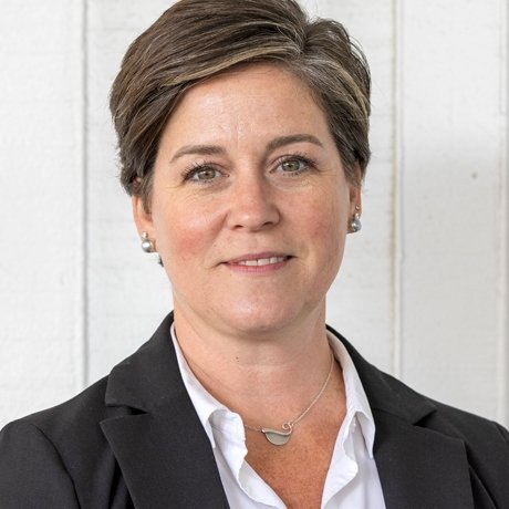 A woman with short brown hair, wearing a black blazer and white blouse, standing against a white wooden background.