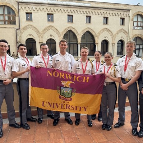 Norwich cadets stand shoulder-to-shoulder holding a Norwich flag, flanked on each end by Norwich faculty. They are standing in a courtyard with a building in the background.