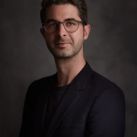 Portrait of an individual with glasses wearing a dark blazer, set against a textured grey background.