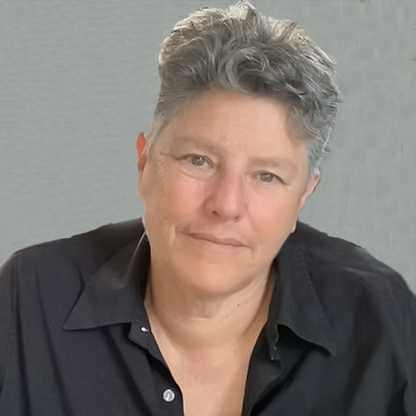 A person with short, wavy gray hair wearing a black button-up shirt, smiling softly, against a soft gray background.