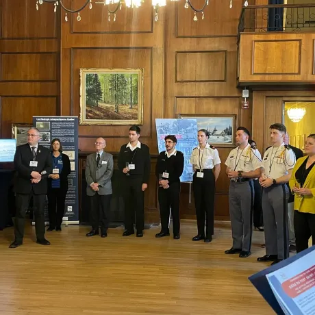 Group of individuals in professional attire standing in a wood-paneled room, with paintings and informational posters on display.