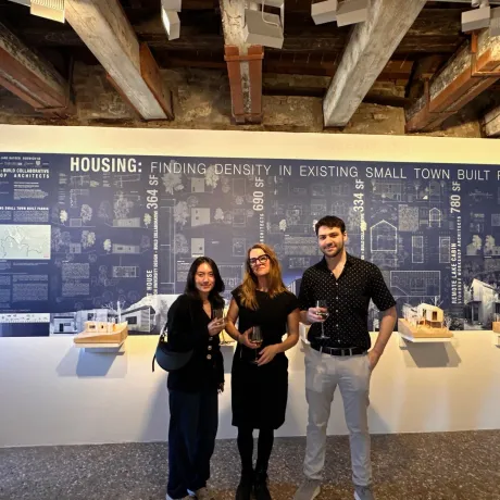 Three individuals holding drinks stand in front of an exhibition panel titled "HOUSING: FINDING DENSITY IN EXISTING SMALL TOWN BUILT FABRIC" at an architectural gallery. They are smiling.