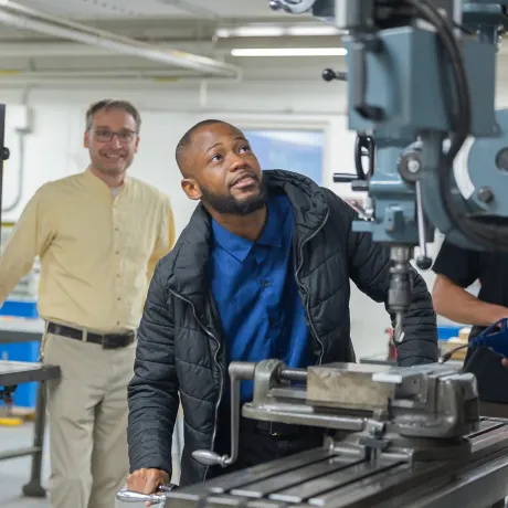 An individual operating a vertical milling machine in an industrial setting while another person observes in the background.
