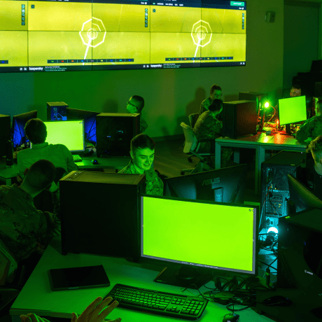 Students in military uniforms working at desktop computers in a green-lit Norwich University cybersecurity lab, with digital displays showing network diagrams.