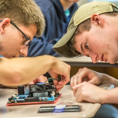 Two Norwich University students work with focused concentration, assembling electronic components on a circuit board at a classroom table.