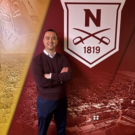 Person standing with arms crossed in front of a Norwich University banner displaying the founding year 1819 and the university emblem.