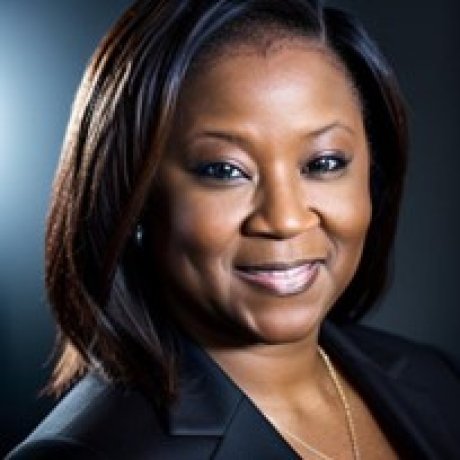 A smiling woman with straight dark hair, wearing a black blazer and gold necklace, posed against a dark background.