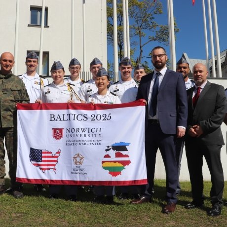 A group photo of the Olmsted trip group, holding a branded Norwich/Olmsted flag in uniform in front of military academy.