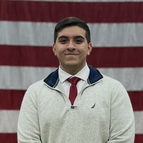 A portrait headshot of Hector Aponte in a sweater, shirt and tie in front of American flag.