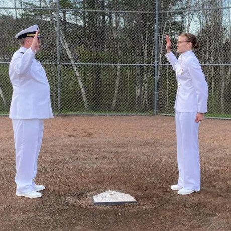 Caitlin Dailey (right) stands next to home plate while her father (left, directly across home plate) swears her in. They are both in full white Navy outfits.