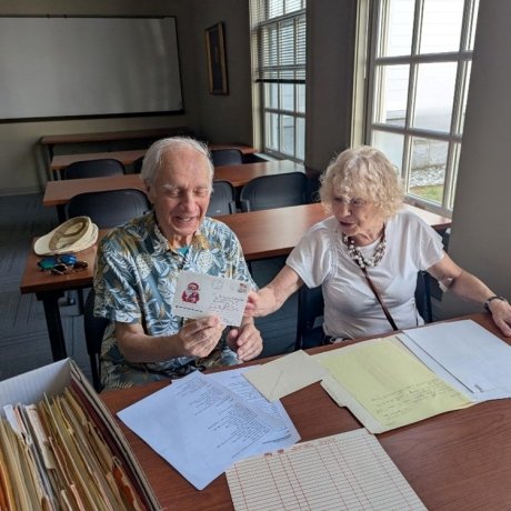 Sergey (left) in a Hawaiian styled shirt holds a letter while sitting at a table, while his wife Anna (right) points at the letter while smiling. Papers are strewn across the table.