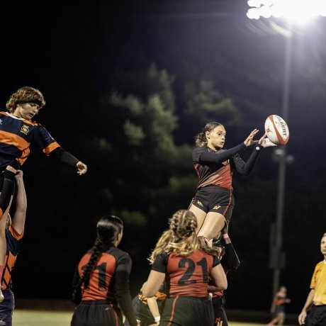 A women's rugby player is lifted into the air while holding the ball during a night match.