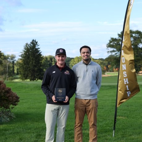 Two people stand on a golf course, one holding a plaque between two gold flags.
