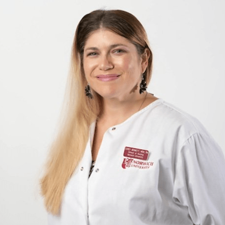 A woman wearing a white lab coat with a Norwich University logo smiles at the camera against a plain white background. She has long light brown hair, partially over one shoulder, and is wearing black earrings.