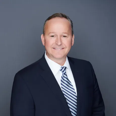 Person wearing a dark suit and striped tie poses against a gray background.