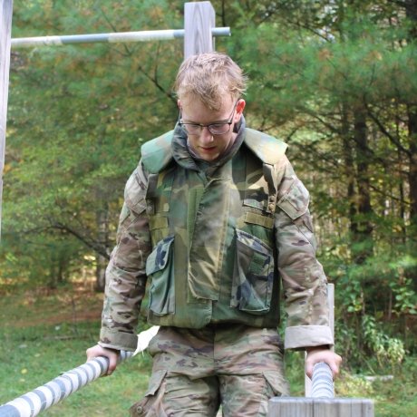 A person in military camouflage uniform performing an exercise on parallel bars in an outdoor obstacle course. The background features a wooded area with green foliage.