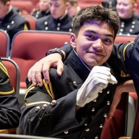 A cadet in a formal military uniform sits in an auditorium seat, smiling at the camera while raising a gloved hand in a friendly gesture