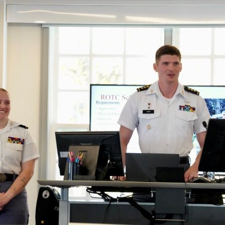 Two cadets in uniform are standing at the front of a classroom giving a presentation. One is speaking while the other smiles and listens. Both are wearing white shirts with rank insignia and ribbons. Behind them, a screen displays a slide with “ROTC Scholarship Requirements” listed. The environment appears professional and academic, with computers, notes, and coffee cups visible on the desks.